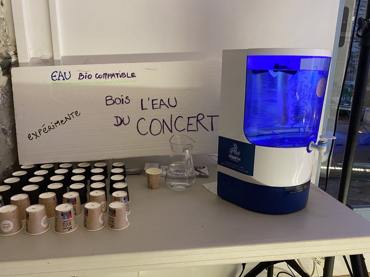 Une fontaine à eau au réservoir éclairé en bleu, posée sur une table à côté d’une carafe et de rangées de gobelets en carton ; derrière, une pancarte manuscrite : « Eau bio compatible — expérimente — Bois l’eau du concert ».
