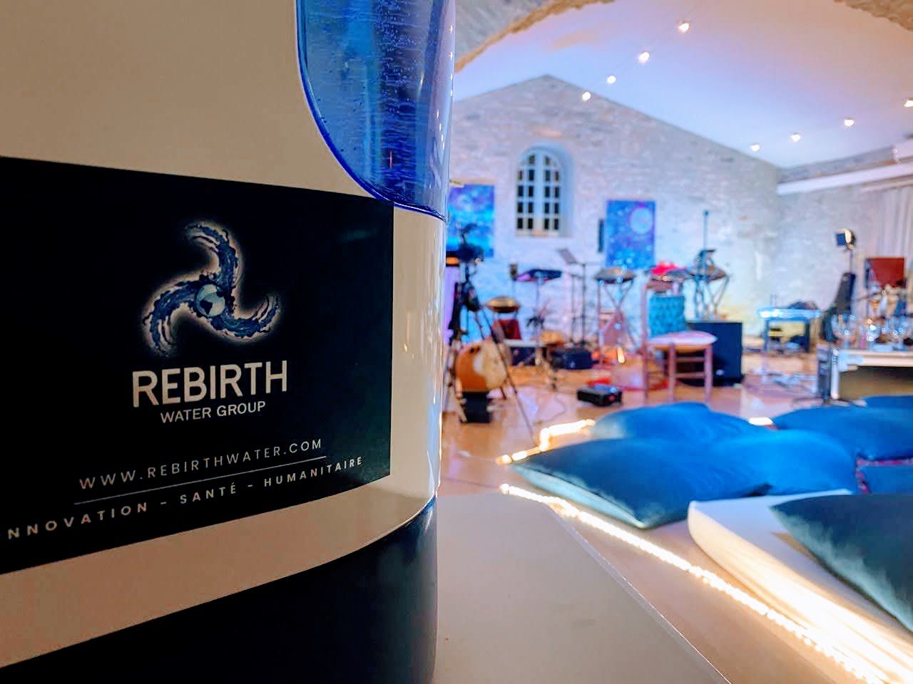 Gros plan sur la fontaine : son étiquette porte le logo Rebirth Water Group ; à l’arrière-plan, une salle de concert voûtée en pierre, les instruments installés et des coussins bleus posés au sol.