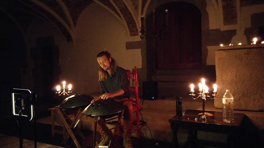 Sous une voûte de pierre éclairée à la bougie, l’artiste assis derrière deux handpans posés sur pieds, entre deux chandeliers allumés.