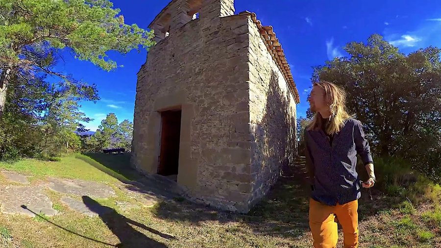 Une petite chapelle de pierre sous un ciel bleu vif, un arbre au-dessus ; l’artiste debout devant la porte, de profil.