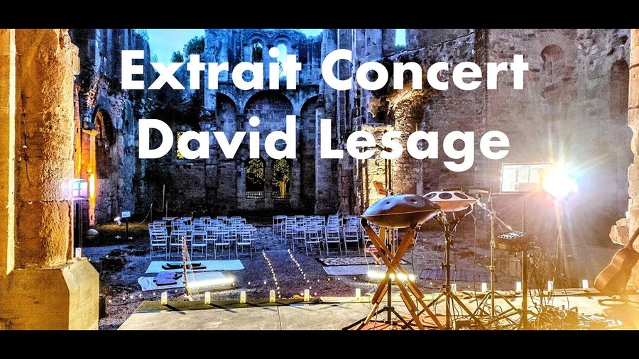 Plateau installé au pied d’un haut mur de pierre éclairé de doré : deux handpans sur pieds au centre, des rangées de chaises vides et un projecteur en contre-jour ; titre incrusté « Extrait Concert David Lesage ».