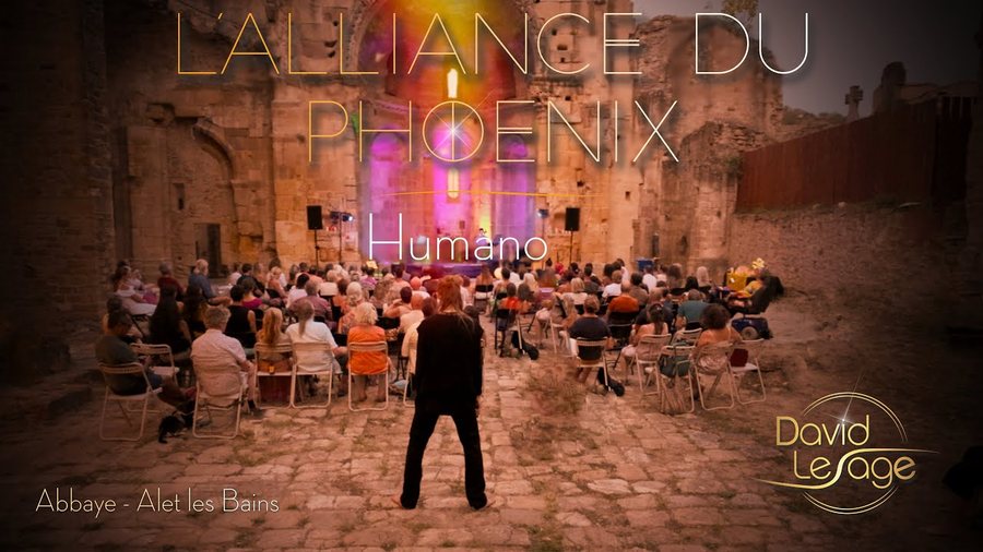 Vue depuis le fond de l’abbaye à ciel ouvert d’Alet-les-Bains : un public nombreux assis sur des chaises entre les murs de pierre, l’artiste seul debout au fond ; titre incrusté « L’Alliance du Phoenix — Humano ».