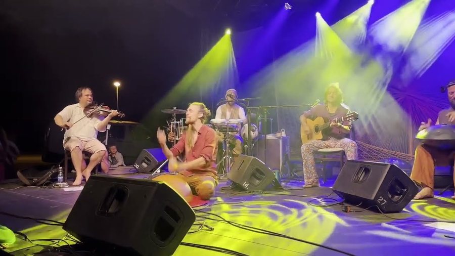 Grande scène de festival dans des faisceaux violets et verts : cinq musiciens assis en arc de cercle, violon, guitare et percussions, des retours de scène au premier plan.