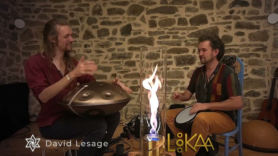 Deux musiciens assis face à face devant un mur de pierre, chacun derrière un handpan acoustique en bronze, une flamme entre eux.