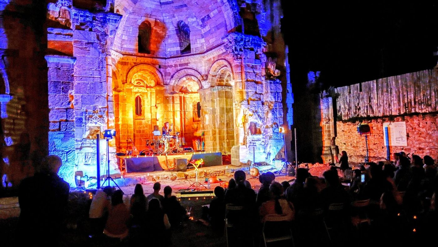 Intérieur d’une abbaye en ruine, à ciel ouvert, éclairé en bleu et en orange ; au fond le plateau et ses instruments, au premier plan un public nombreux assis face à la scène.
