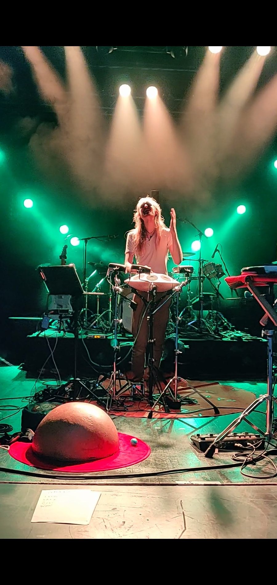 David Lesage seul au centre d’une grande scène de salle, la tête levée en train de chanter et une main en l’air, derrière son Neotone — le handpan électronique à coque claire — posé sur un pied ; autour de lui son installation — pupitre, ordinateur, claviers, batterie — dans des faisceaux de lumière verte et blanche et de la fumée ; au premier plan, sa calebasse posée sur un tapis rond rouge.