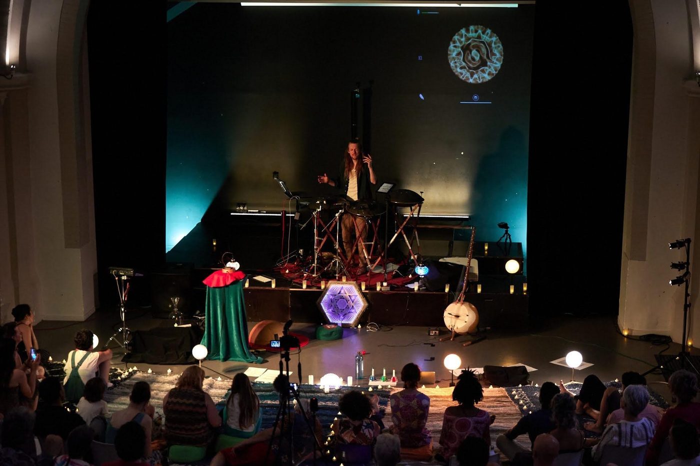 Vue d’ensemble d’une église transformée en salle de concert : David Lesage seul sur l’estrade au milieu de ses instruments, une figure cymatique projetée au-dessus de lui, et le public assis au sol sur des tapis parmi des bougies.