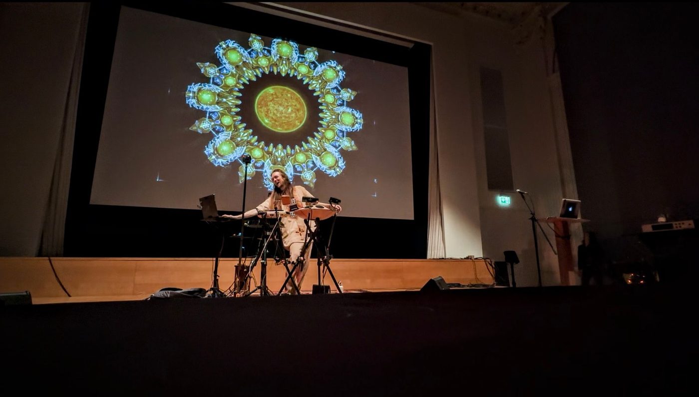 David Lesage seul au centre d’un plateau, derrière ses instruments, devant un très grand écran où est projetée une figure cymatique verte et dorée en forme de fleur.