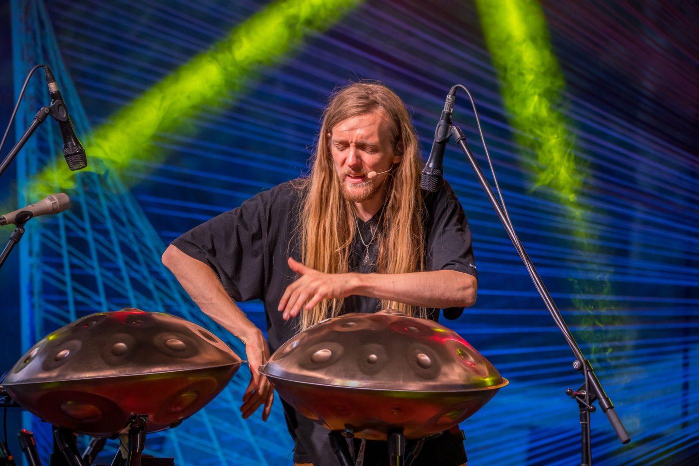 David Lesage seul sur scène, buste et visage de face, un micro-serre-tête au visage, penché entre deux handpans acoustiques Yishama en acier bruni posés sur pieds ; ses deux mains sont en mouvement au-dessus de celui de droite, dont les creux sont nettement visibles. Deux micros sur pieds l’encadrent ; derrière lui, de larges faisceaux de lumière verte croisent un fond bleu tendu de fils.