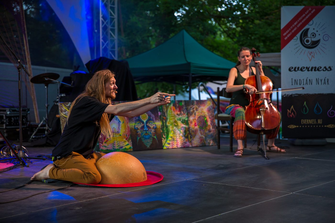 David Lesage assis sur ses talons au bord d’un plateau de festival, derrière une grande calebasse claire posée sur un tapis rond rouge, pieds nus, en tee-shirt noir et pantalon ocre : les deux bras tendus devant lui et l’index pointé vers le public hors champ, la bouche ouverte en train de chanter. Derrière lui, des panneaux peints de couleurs vives, la violoncelliste invitée Valeria Pribay assise sur une chaise qui joue, et deux banderoles du festival Everness ; au fond, une tente de plein air et des arbres.