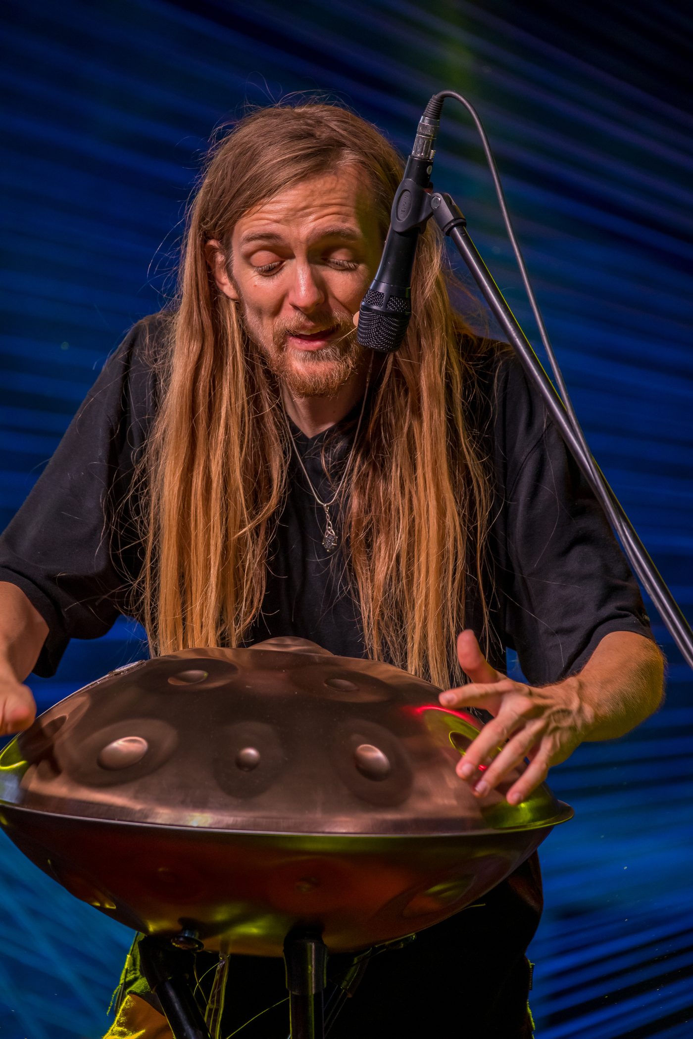 Gros plan vertical : David Lesage penché sur un handpan acoustique Yishama en acier bruni posé sur pied, les deux mains sur l’instrument dont les creux et le dôme central sont nettement visibles ; cheveux longs retombant sur le côté, micro-serre-tête et micro de scène près du visage, regard baissé sur l’instrument, sur un fond bleu strié de faisceaux verts.