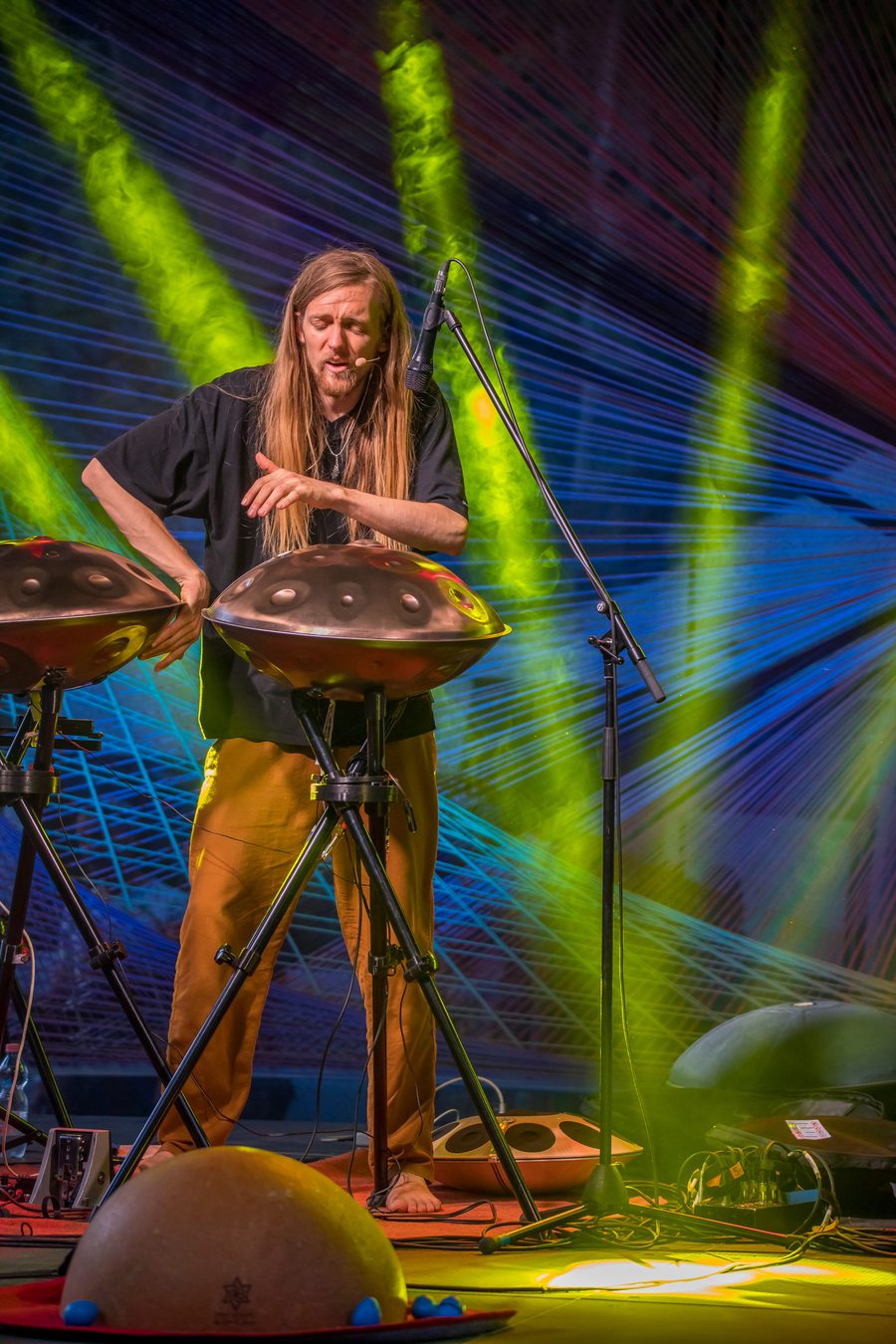 Plan vertical en pied : David Lesage debout et pieds nus sur une scène de festival, en tee-shirt noir et pantalon ocre, les mains au-dessus de deux handpans acoustiques Yishama posés sur pieds devant lui, un micro-serre-tête au visage et un micro sur pied à sa droite. Au premier plan au sol, sa grande calebasse claire posée sur un tapis rond, son Neotone à coque claire et un boîtier de commande à même le sol, câbles apparents ; derrière lui, des faisceaux de lumière verte et bleue sur un fond tendu de fils.