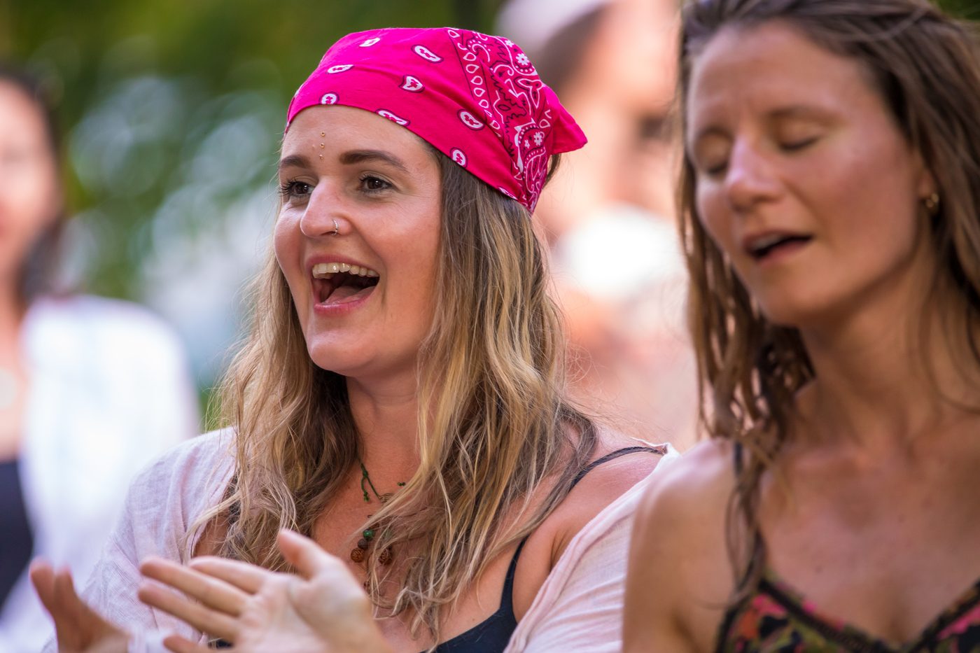 Gros plan de deux spectatrices côte à côte dans le public, en plein air : à gauche une femme au foulard rose vif chante à pleine bouche, les mains levées devant elle ; à droite, une autre femme chante les yeux fermés. Fond de feuillage flou.