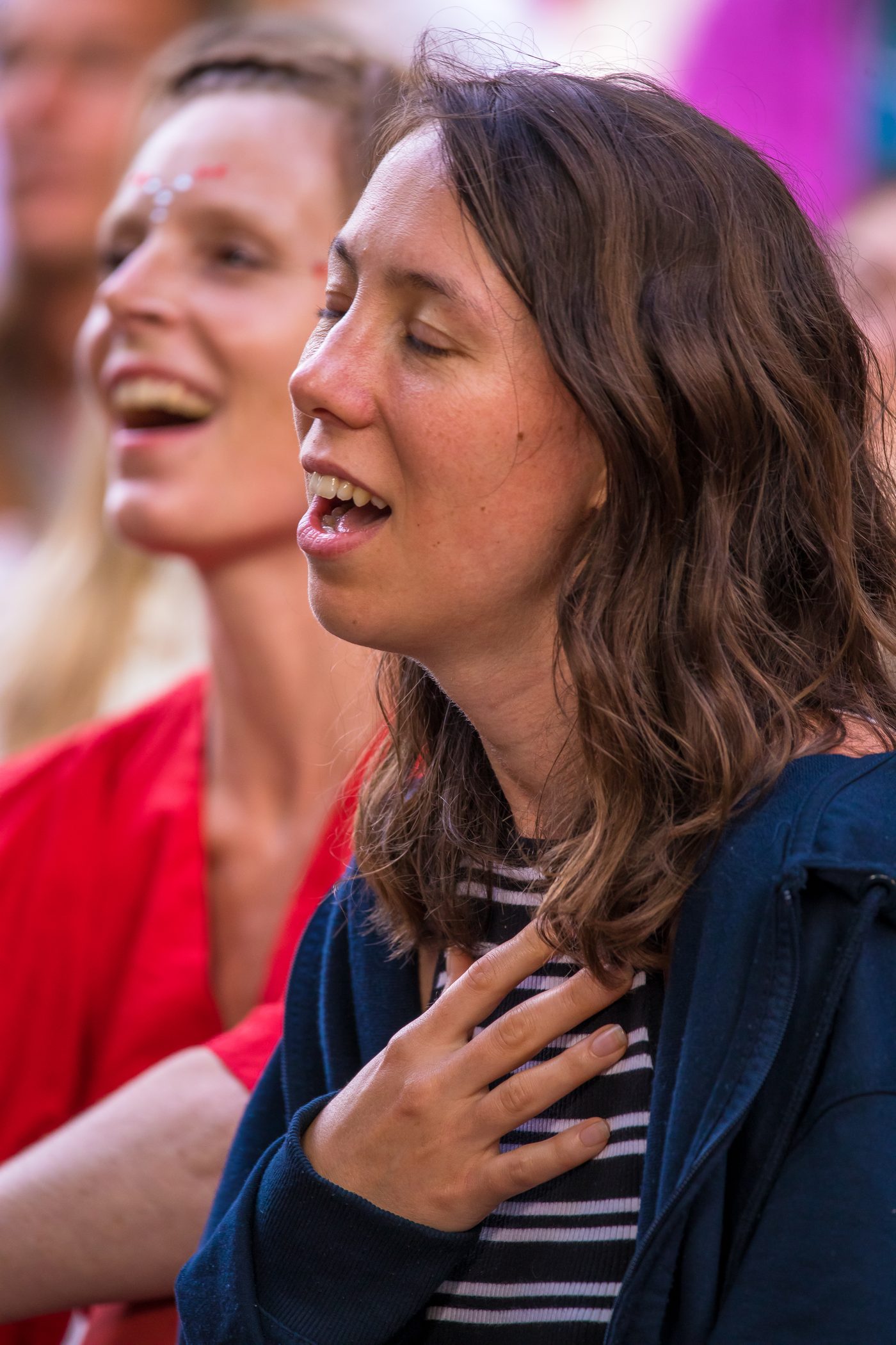 Gros plan vertical d’une spectatrice au milieu du public, en plein air : les yeux fermés, la bouche ouverte en train de chanter, une main à plat sur le sternum ; derrière elle, une autre femme chante aussi, en veste rouge. Fond de public flou.