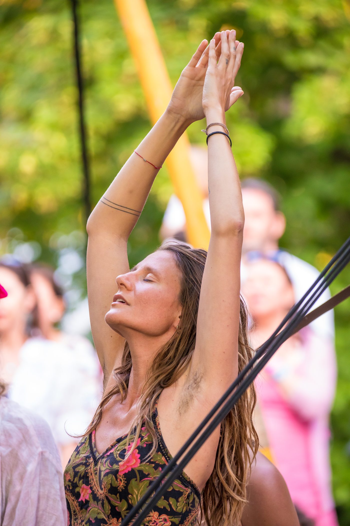 Gros plan vertical d’une spectatrice debout dans le public : les deux mains jointes très haut au-dessus de la tête, le visage levé et les yeux fermés ; derrière elle, d’autres personnes debout et du feuillage vert.