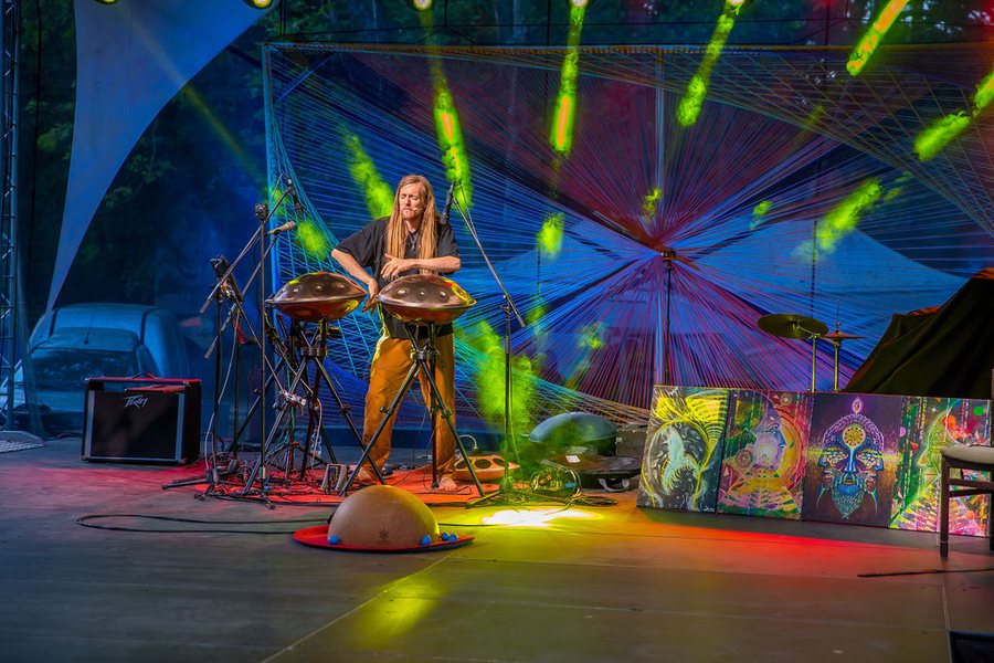 David Lesage seul debout au centre d’une grande scène en plein air, les mains sur deux handpans acoustiques Yishama posés sur pieds ; derrière lui une immense toile de fils tendus multicolores traversée de faisceaux de lumière verte, devant lui une calebasse sur un tapis rond rouge.