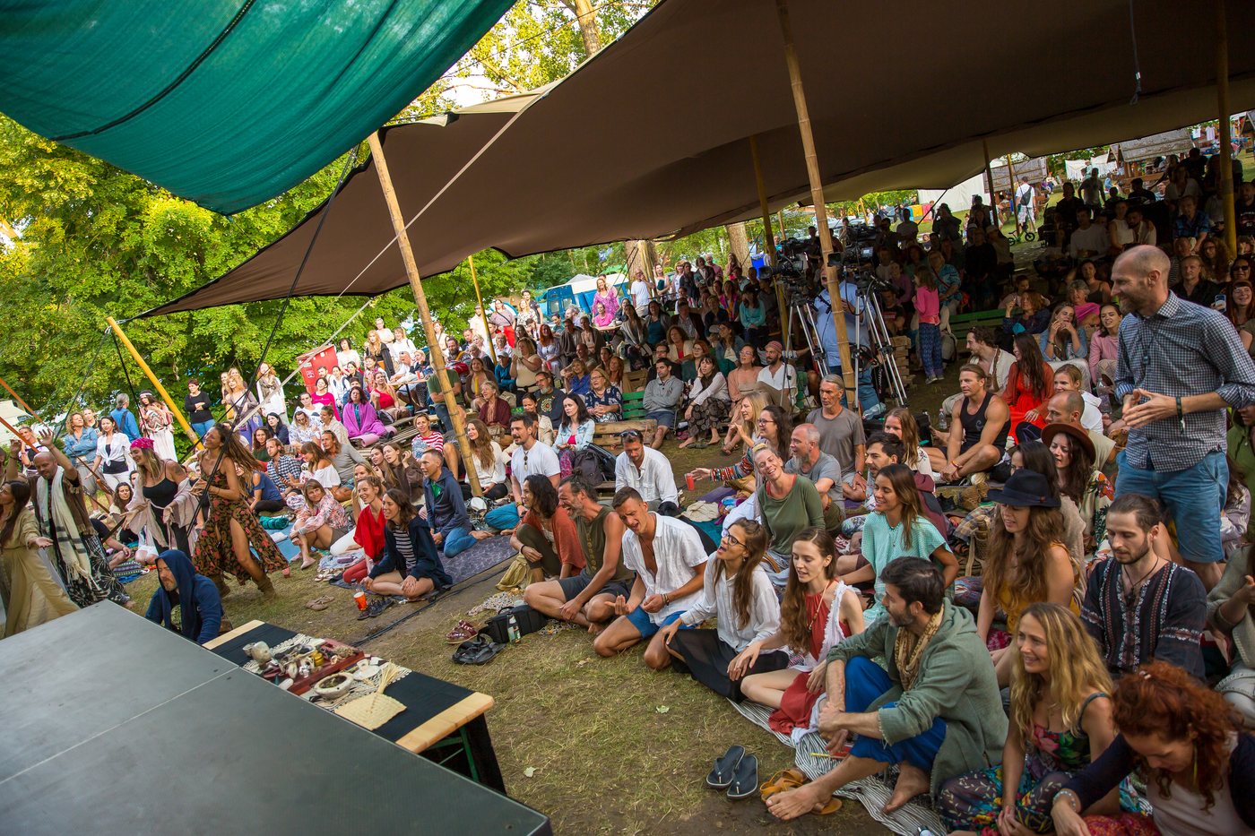 Vue prise depuis le bord du plateau : un public très nombreux assis dans l’herbe et sur des tapis, sous de grandes voiles d’ombrage tendues entre des perches, en plein air ; au fond, des rangs debout et les stands du festival sous les arbres.