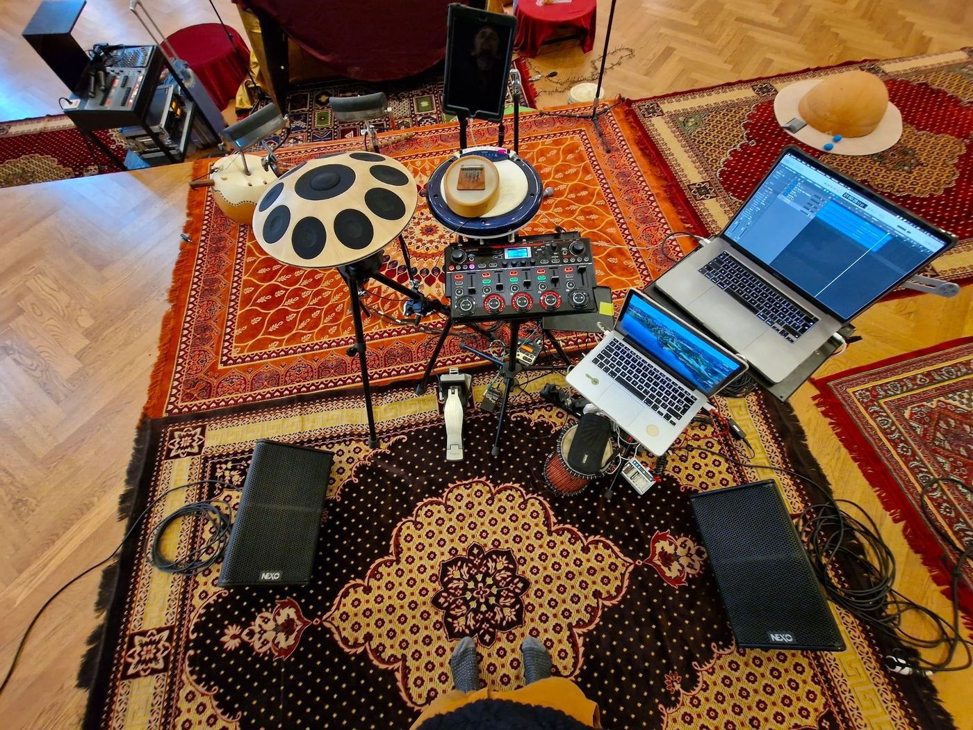 Vue plongeante du setup installé sur deux tapis persans : un handpan électronique et un pad de percussion sur pieds, une loop station, deux ordinateurs portables ouverts, une tablette, deux enceintes de retour posées au sol et une calebasse.