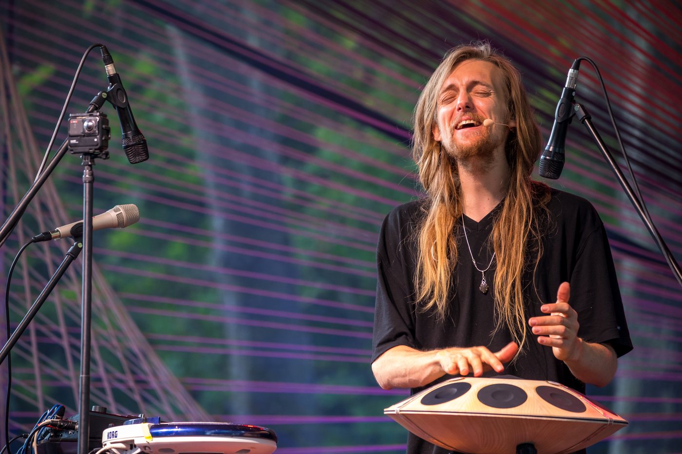 David Lesage seul sur scène, en plan rapproché, les yeux fermés en train de chanter dans un micro serre-tête, les deux mains posées sur un Neotone — le handpan électronique, coque de bois clair et pastilles sombres. Deux micros sur pied l’encadrent, un contrôleur Korg est posé au premier plan ; en fond, un décor de fils tendus violets, verts et rouges devant de la végétation.
