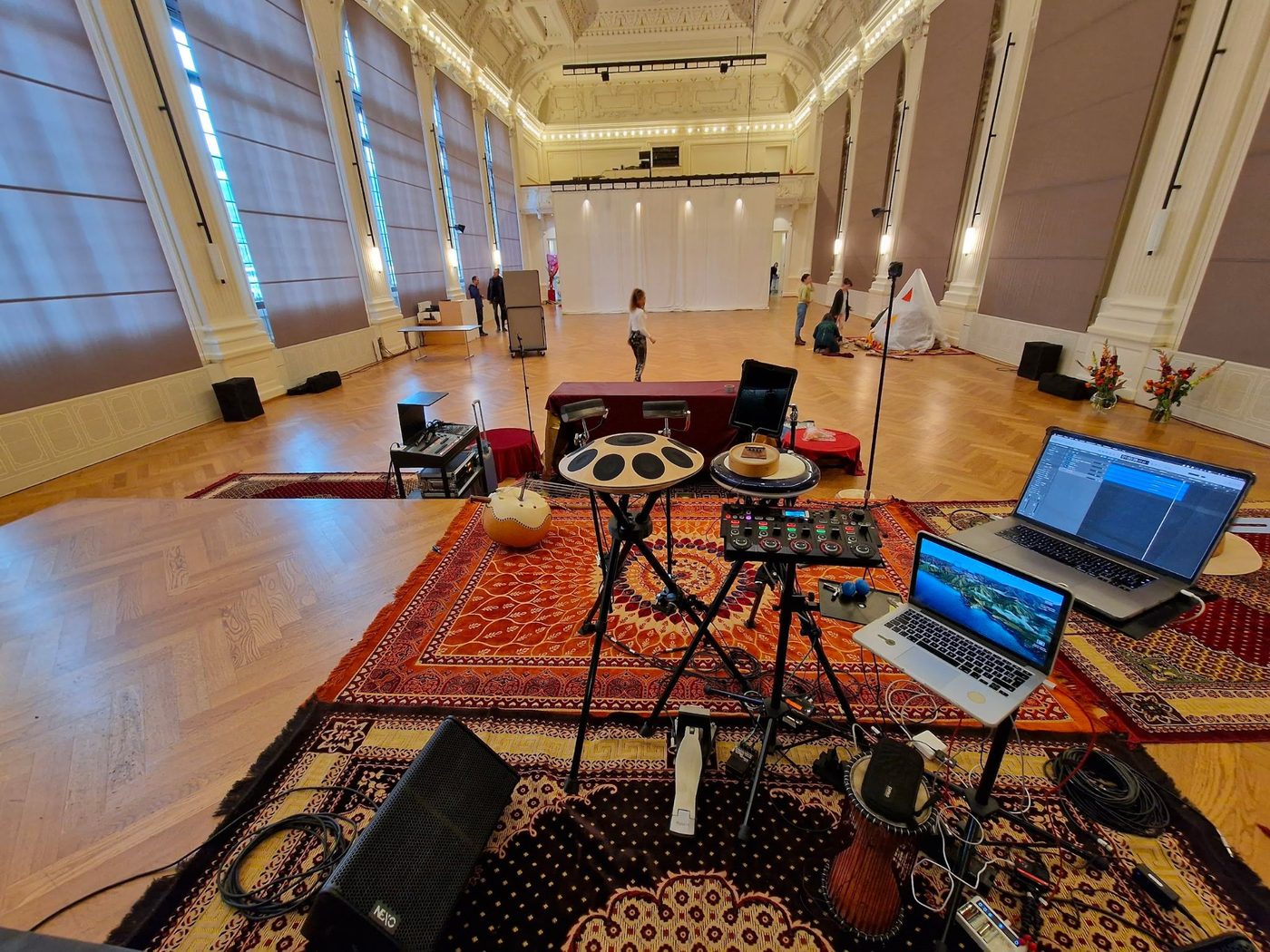 Grande salle claire à colonnes et hautes fenêtres, vue depuis la scène : au premier plan le plateau installé sur des tapis avec handpans, ordinateurs, calebasse et enceintes de retour, et au fond le parquet vide de la salle.