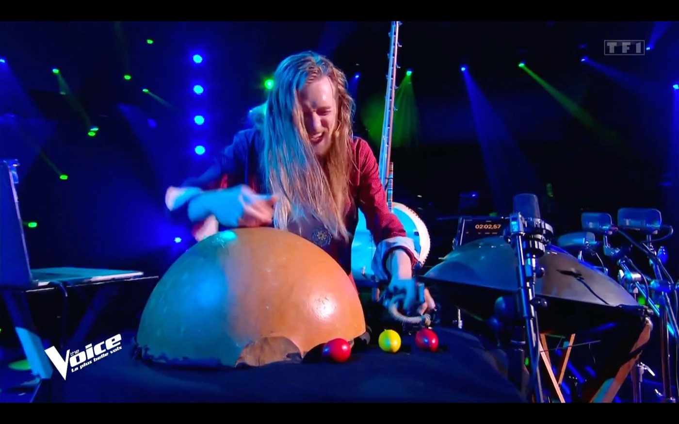David Lesage seul sur le plateau de The Voice, penché sur une grande calebasse posée devant lui, les deux mains dessus, dans une lumière bleue ; à sa droite ses handpans sur pieds. Logos The Voice et TF1 incrustés sur l’image.