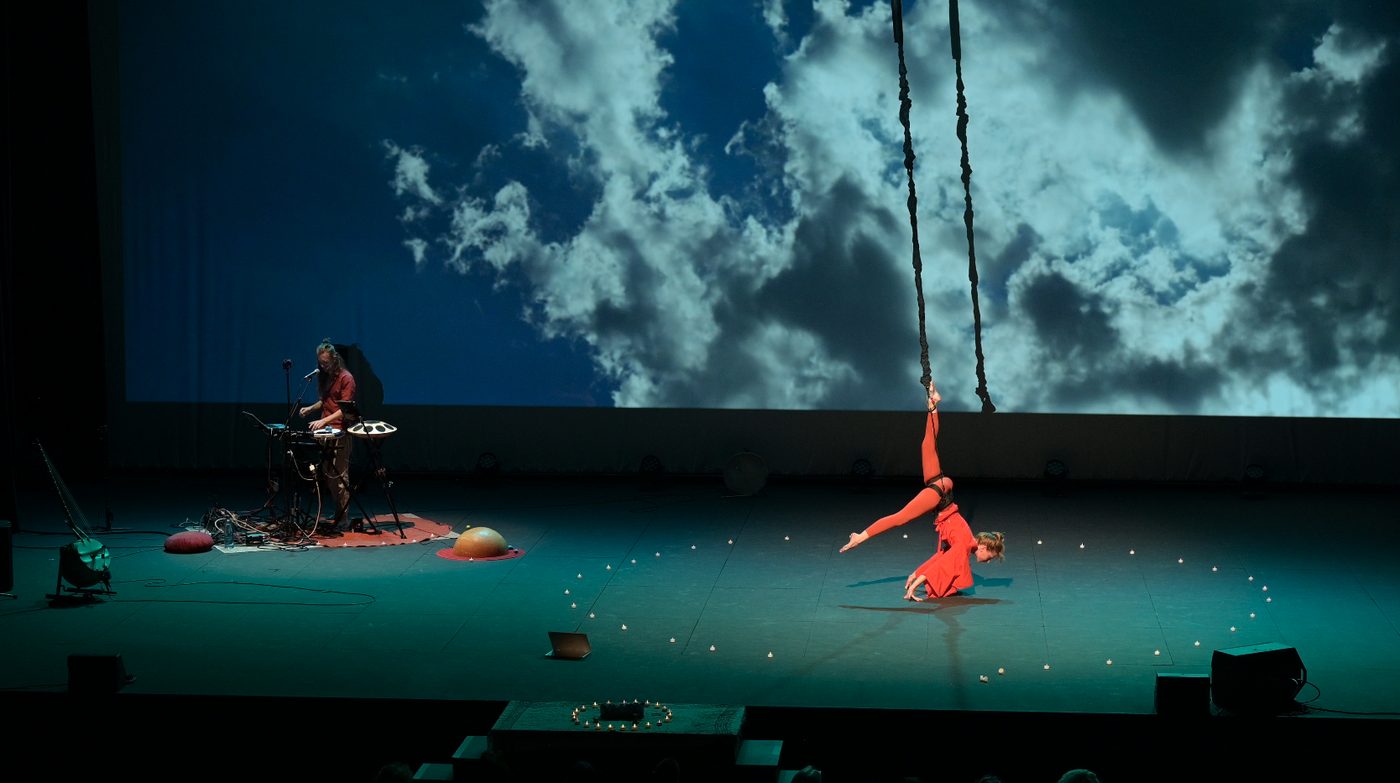 Grande scène : à gauche David Lesage à son handpan sur un tapis, à droite Iris Chasles en tenue rouge suspendue à un élastique, en équilibre au sol, devant une vidéoprojection de nuages et de ciel bleu.