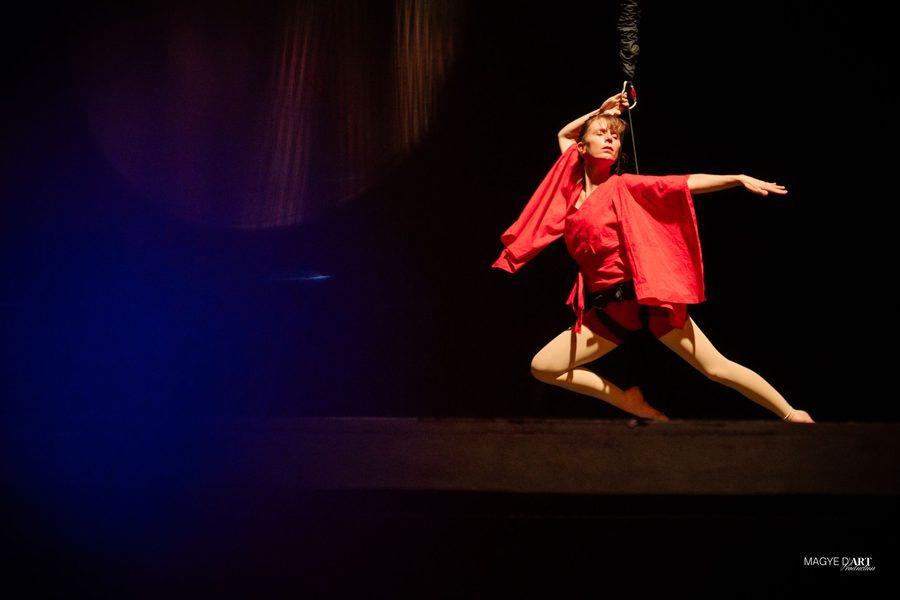 Iris Chasles, en tunique rouge et en grande fente sur le plateau du Grand Rex, tenue par son baudrier à l’élastique qui descend des cintres ; derrière elle, un fond bleu profond.