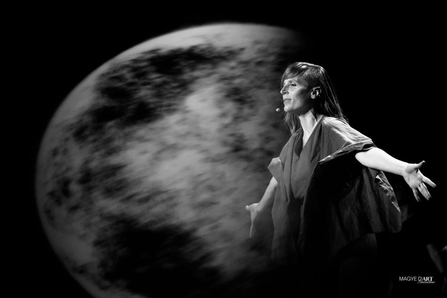 Photo en noir et blanc : Iris Chasles, micro-casque au visage et bras ouverts, se détache devant l’immense lune projetée sur l’écran du Grand Rex.