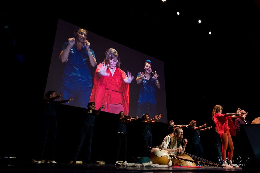 Sur le plateau du Grand Rex, Iris Chasles en rouge guide les gestes de la salle, tandis que David Lesage, assis derrière sa calebasse, impulse le rythme pour que tout le monde soit ensemble dans le même geste ; autour d’eux une dizaine d’artistes en tenue sombre, et au-dessus l’écran géant qui les reprend en gros plan.