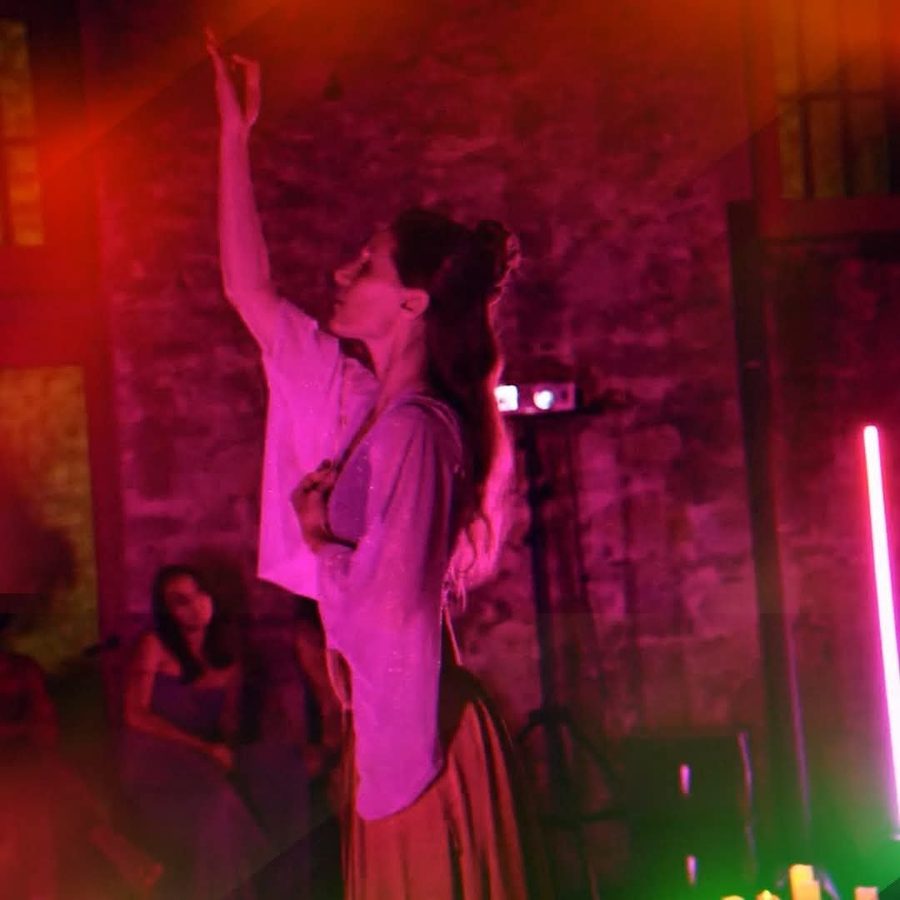 Iris Chasles en danse de tournoiement, un bras levé vers le haut, le visage tourné vers sa main, dans une lumière rose et rouge devant un mur de pierre.