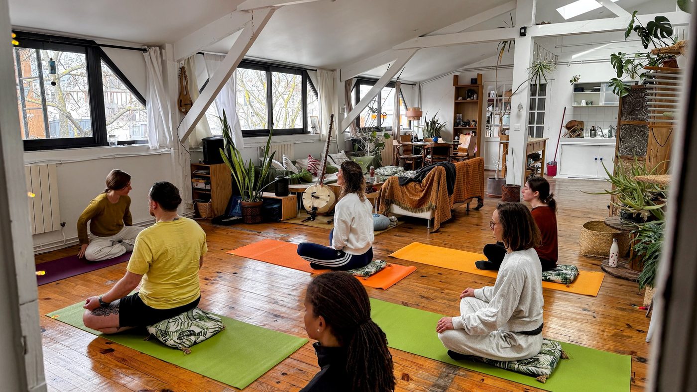 Atelier de yoga au Nid : un groupe assis en méditation sur des tapis colorés, face à l’intervenante, dans une grande pièce mansardée aux fenêtres claires et aux plantes