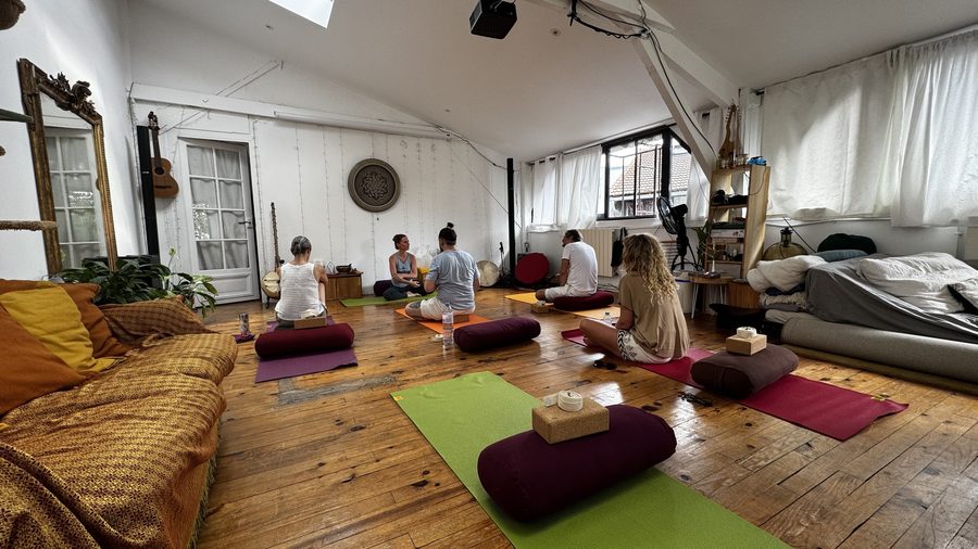 Plusieurs personnes assises jambes croisées sur des tapis de yoga verts et rouges, avec bolsters et briques, dans la grande pièce mansardée au parquet ancien