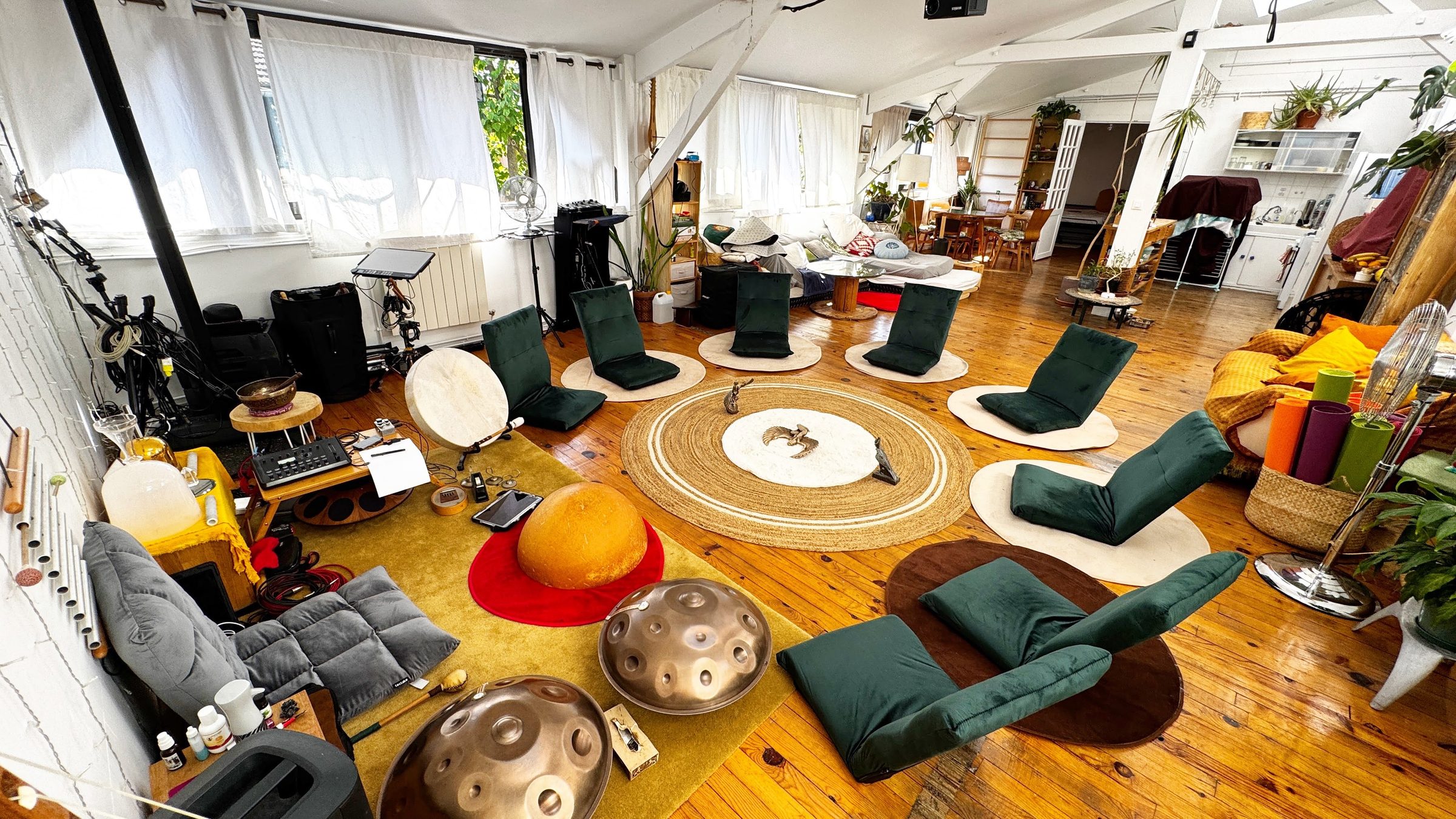 Le salon du Nid : des sièges bas et des coussins disposés en cercle sur le parquet autour d’un tapis rond, deux handpans posés au sol au premier plan, de grandes verrières et des plantes.