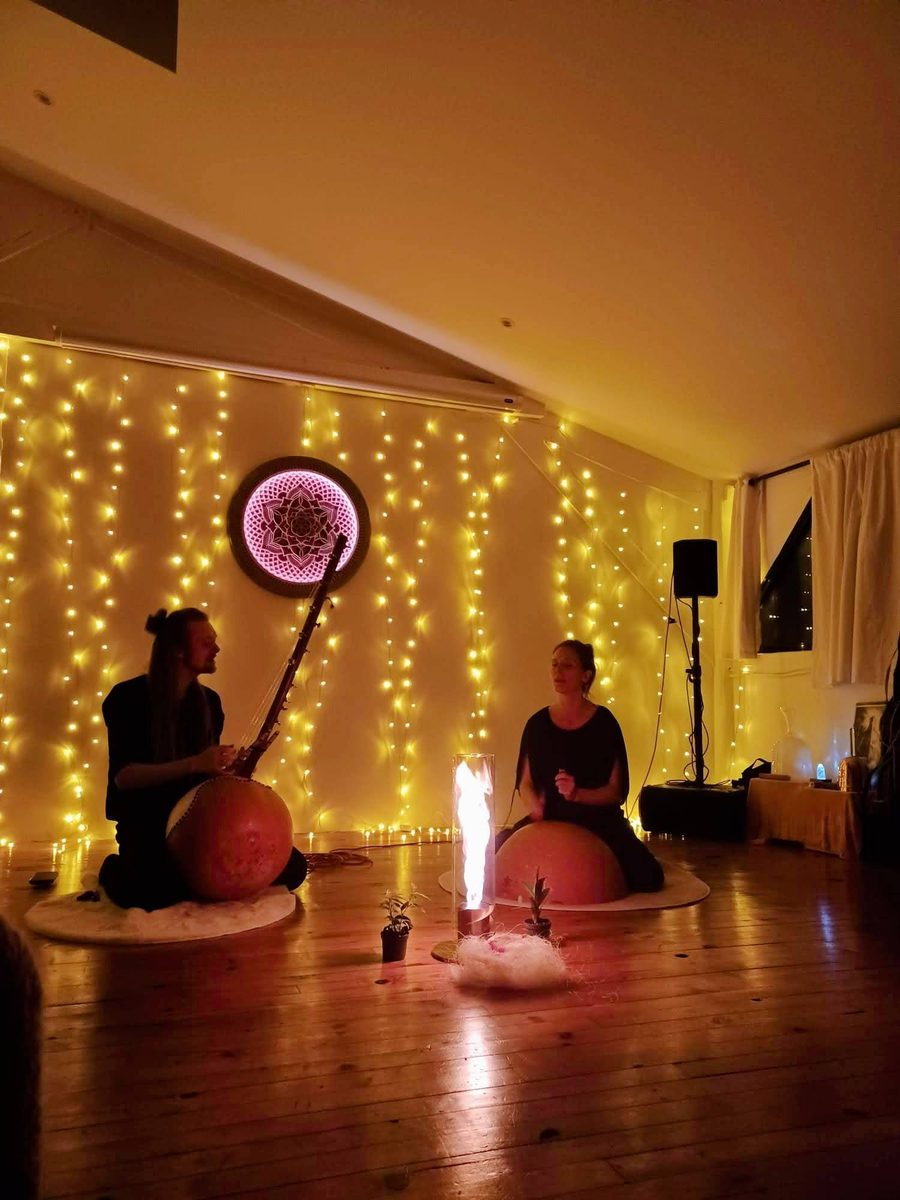 Deux musiciens assis au sol jouent de la calebasse devant un mur de guirlandes lumineuses et un mandala lumineux, une lampe à flamme posée entre eux