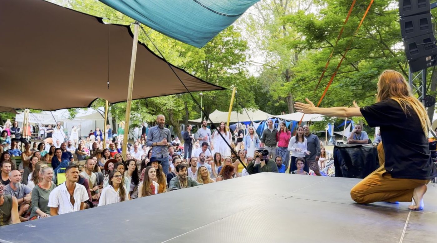 David Lesage seul sur une grande scène de festival en plein air, de dos, les bras ouverts vers un public nombreux assis et debout sous les arbres.