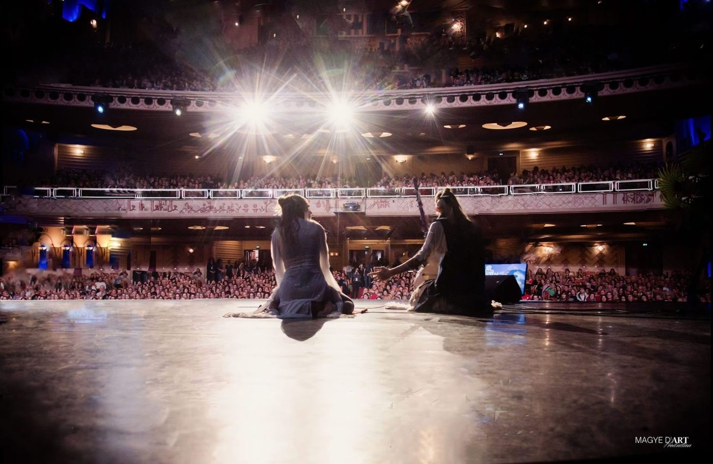 Vue de scène du Grand Rex : deux artistes assis au sol au centre du plateau, face à une salle comble sur deux niveaux, dans les faisceaux de deux projecteurs en contre-jour.