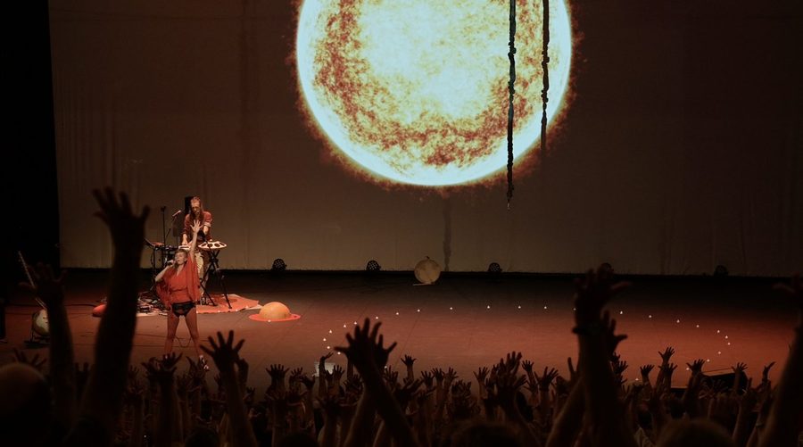 Vue depuis le public d’une grande salle : sur scène, une immense vidéoprojection de soleil orange, deux artistes minuscules devant leurs instruments, et au premier plan une forêt de bras levés.
