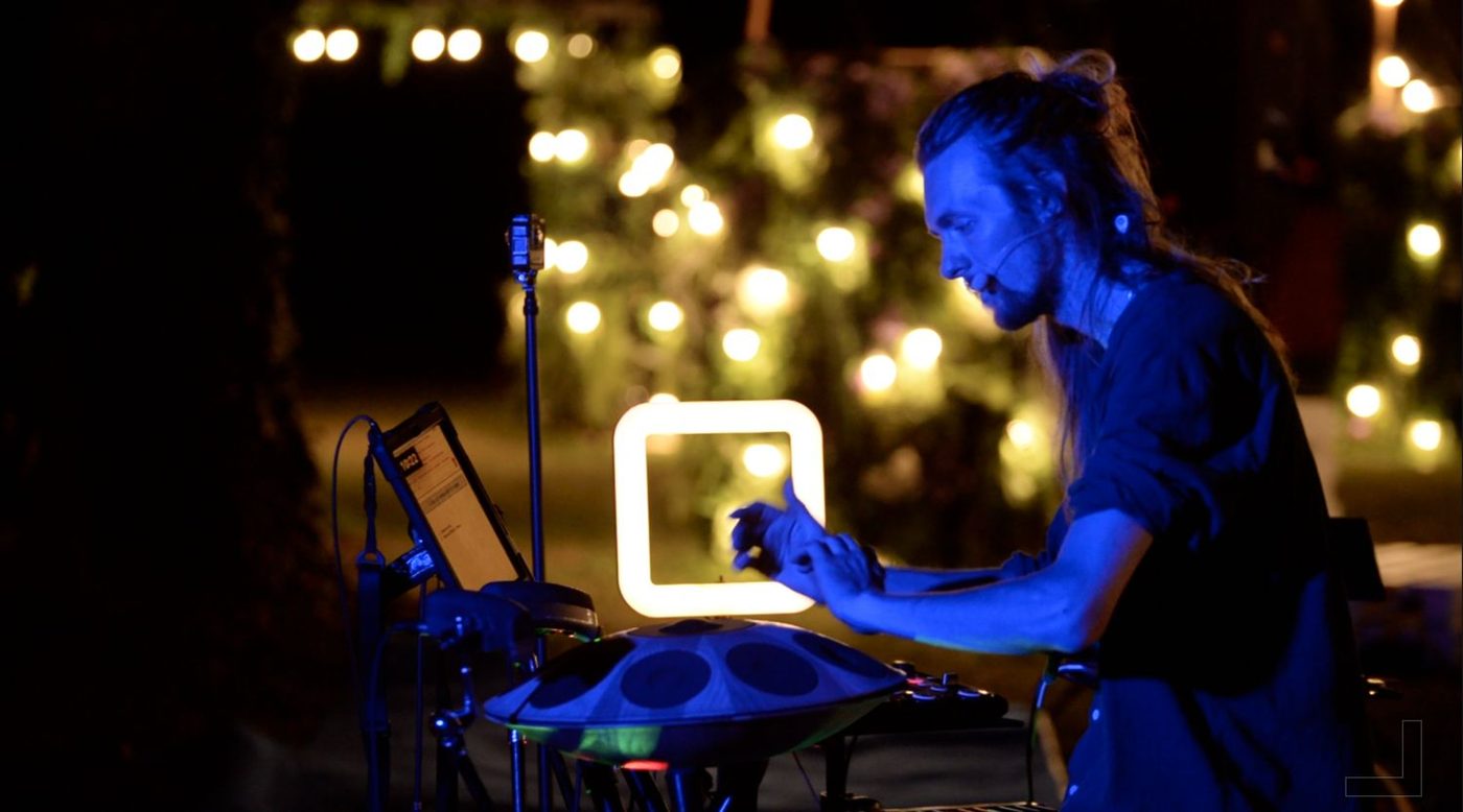 David Lesage seul en concert, penché sur son handpan, éclairé en bleu ; à côté de lui un contrôleur électronique et un micro sur pied, et derrière lui des guirlandes lumineuses dans la nuit.