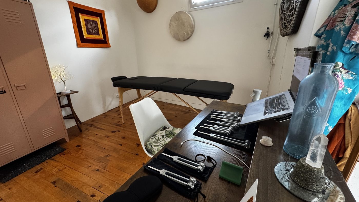 Espace de soin : une table de massage noire dressée près de la fenêtre et, au premier plan, une table de bois où sont alignés plusieurs jeux de diapasons thérapeutiques, à côté d’un grand cristal de quartz.