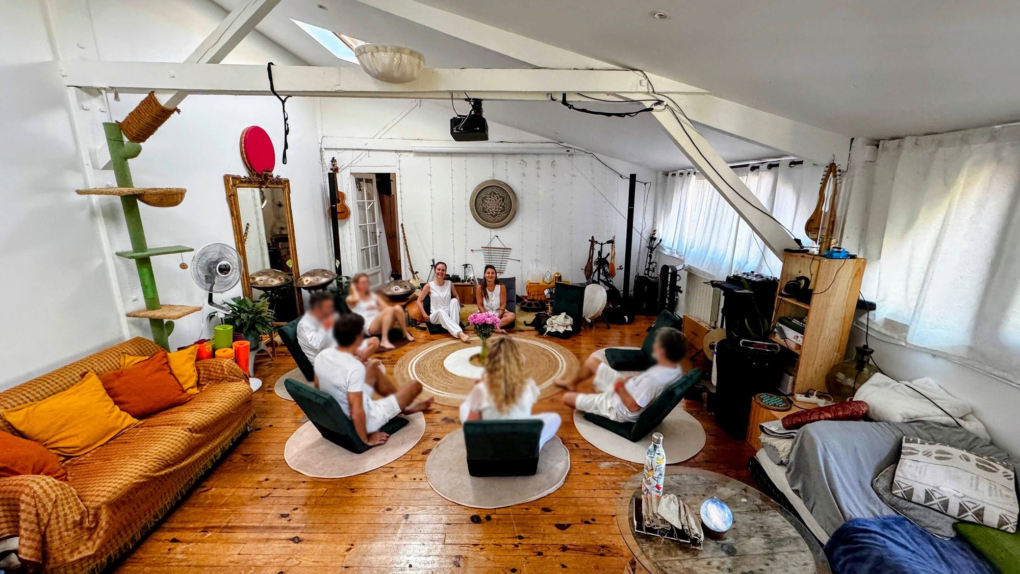Cercle au Nid : une dizaine de personnes vêtues de blanc, assises sur des sièges bas et des coussins autour d’un tapis rond fleuri, deux intervenantes au fond, et des instruments posés au sol — handpans, gong et harpe.